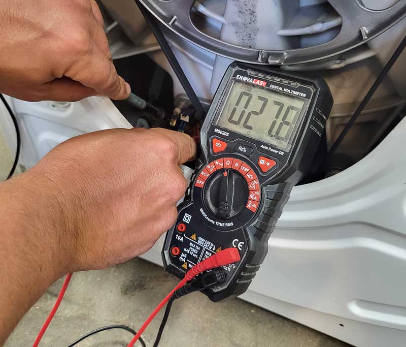 Elektromessgerät klemmt an einer Waschmaschine, zwei Hände halten Teile des Messgeräts