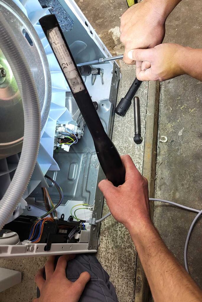 Zwei Techniker schrauben an der Rückseite eines Waschtrockners, einer hält ein Licht