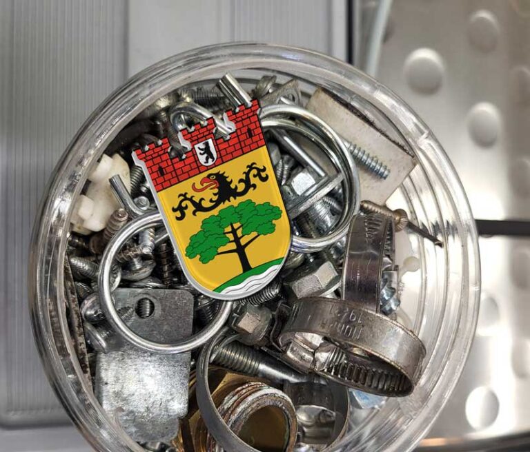 Glas voller Schrauben auf dem ein metallenes Wappen des Bezirks Steglitz-Zehlendorf liegt