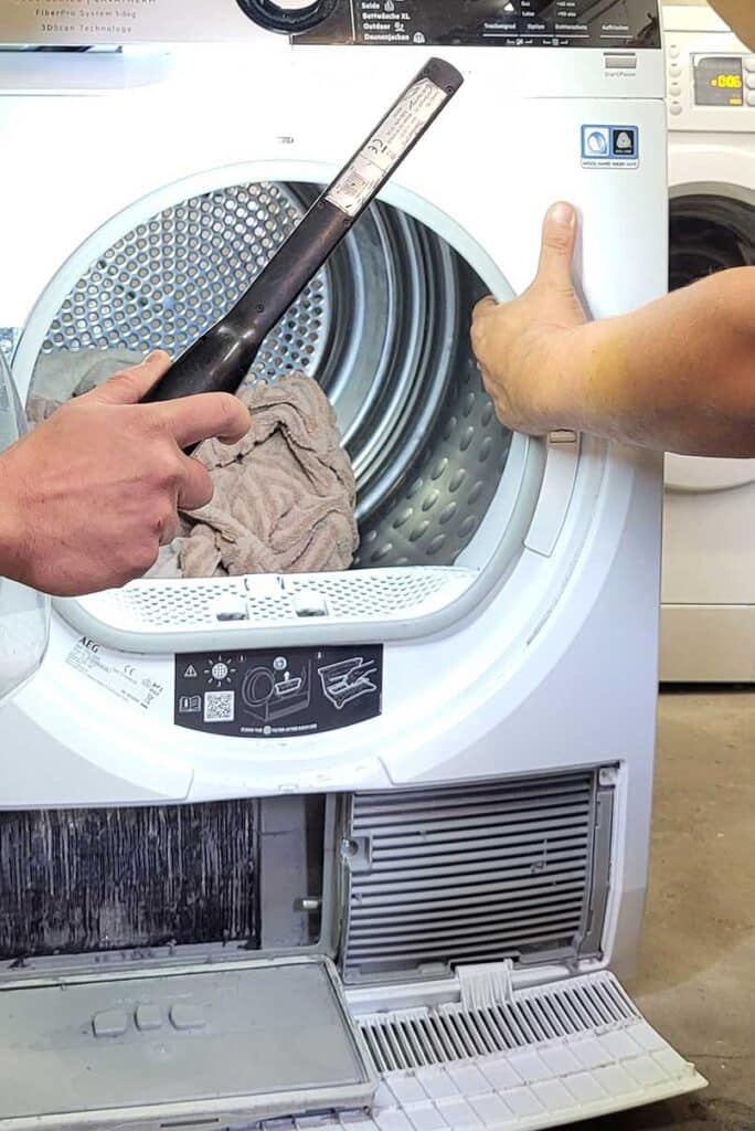 Nahaufnahme eines geöffneten Wäschetrockners, eine Hand greift rein, eine andere Hand hält eine Lampe