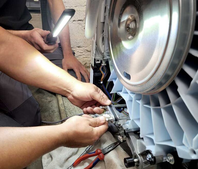 Reparatur einer geöffneten Waschmaschine, zwei Hände montieren ein neues Heizelement, eine andere Hand hält eine Lampe