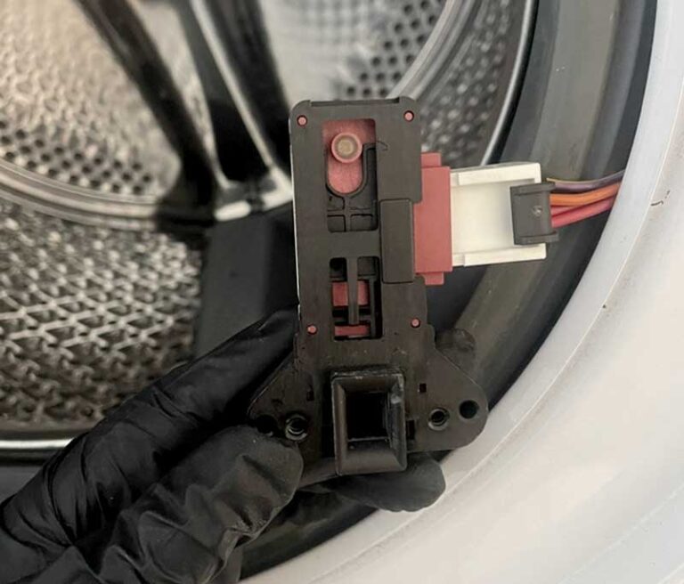Nahaufnahme einer Hand, die eine ausgebaute Türverriegelung eines Waschtrockners vor dem Gerät hält