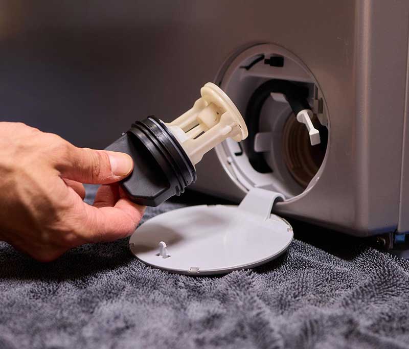 Hand hält Flusensieb vor eine geöffneten Serviceklappe einer Waschmaschine