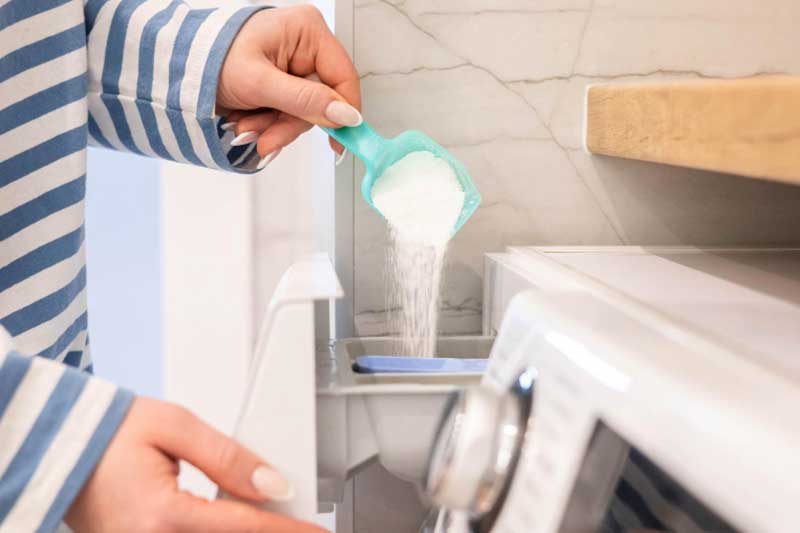Eine Hand füllt Pulver mit einem Dosierlöffel in den geöfneten Waschmaschinedosierkasten