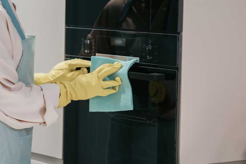 Eine Person mit gelben Schutzhandschuhen reinigt die Glasfront eines schwarzen Einbaubackofens mit einem Mikrofasertuch.