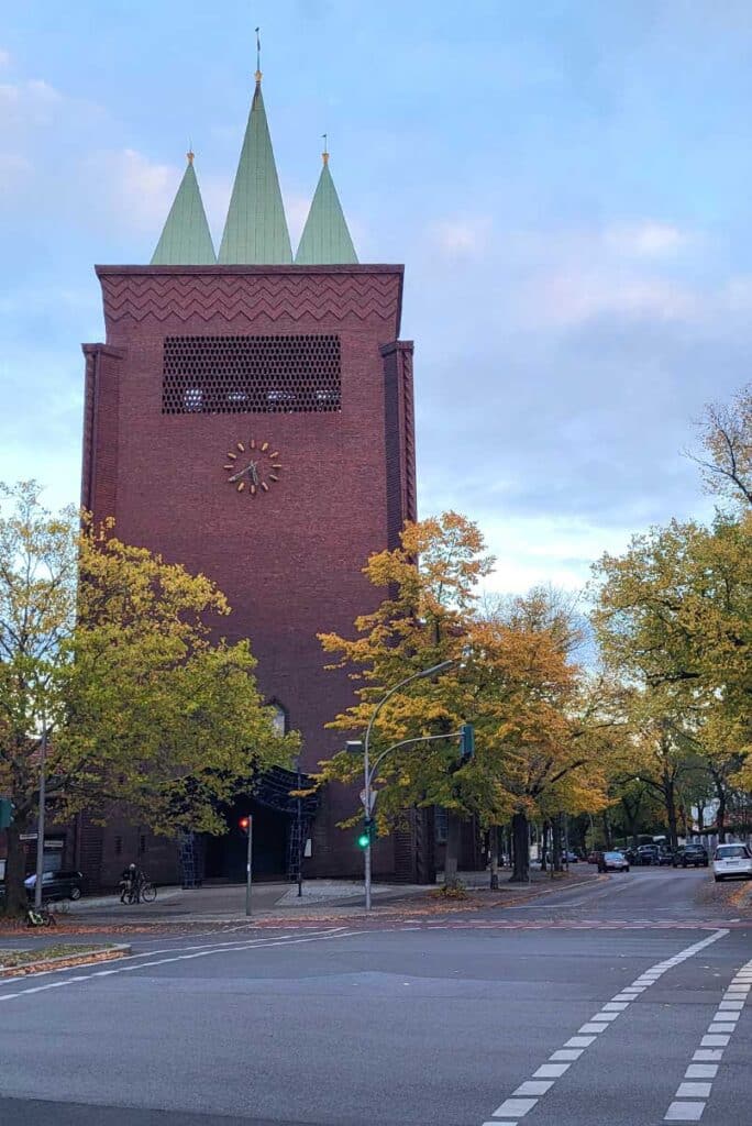 Backsteinturm der evangelischen Kreuzkirche in Berlin-Schmargendorf mit Turmuhr und drei charakteristischen grünen Kupferspitzen, eingerahmt von herbstlichen Bäumen an einer Straßenkreuzung.