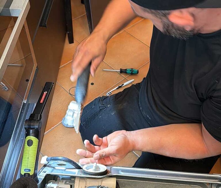 Ein Handwerker kniet auf Küchenfliesen und reinigt mit einem Bürstenaufsatz den Pumpensumpf bzw. Filterbereich eines Haushaltsgeräts neben Arbeitsleuchte und Schraubendreher.
