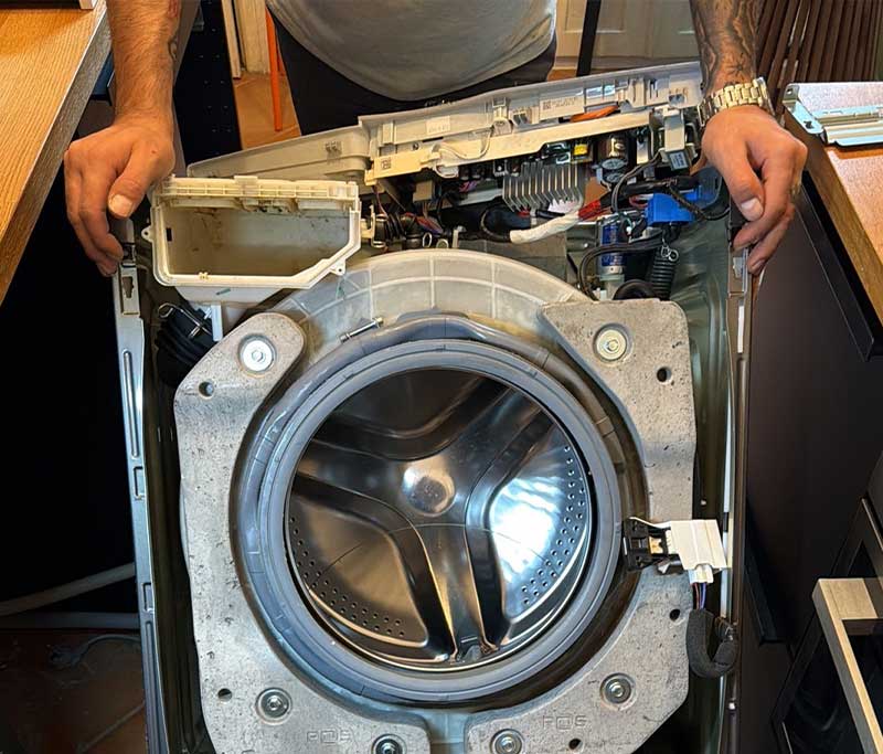 Ein Techniker hält das geöffnete Gehäuse einer freigelegten Waschmaschine mit Blick auf die Edelstahltrommel, das Betongewicht und die obere Steuerelektronik.