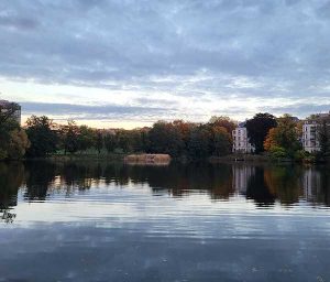 Halensee in Wilmersdorf mit spiegelnder Wasseroberfläche, umgeben von herbstlich gefärbten Bäumen und Wohnhäusern unter bewölktem Abendhimmel.
