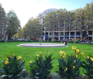 Der gepflegte grüne Prager Platz mit rundem Steinbrunnen und gelben Blumenbeeten vor einem modernen Wohn- und Geschäftshauskomplex.